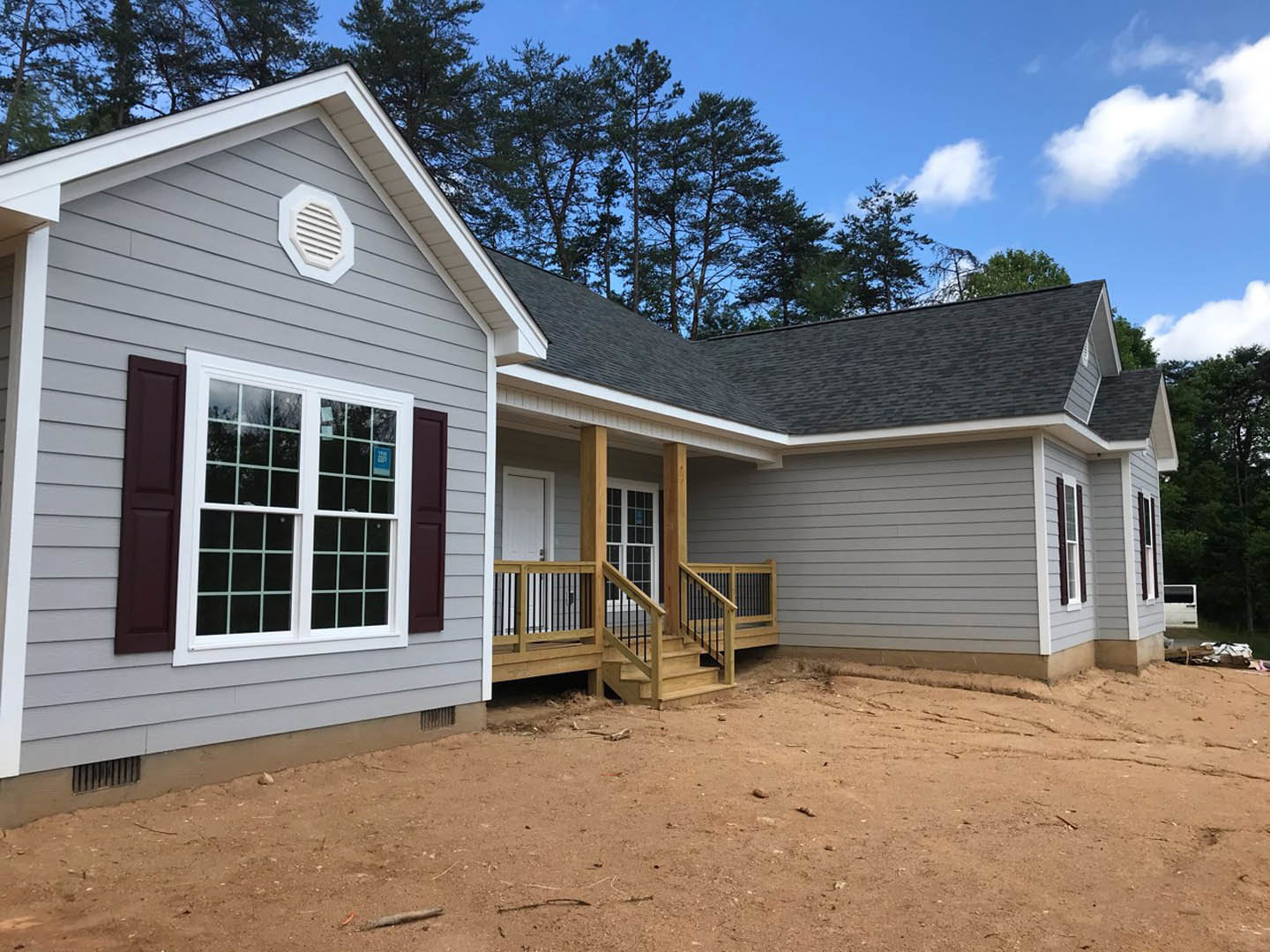 Two-story home with grey siding, white trim, covered porch, wooden stairs leading to entry, large windows, white vent, dirt yard, and trees in background.
