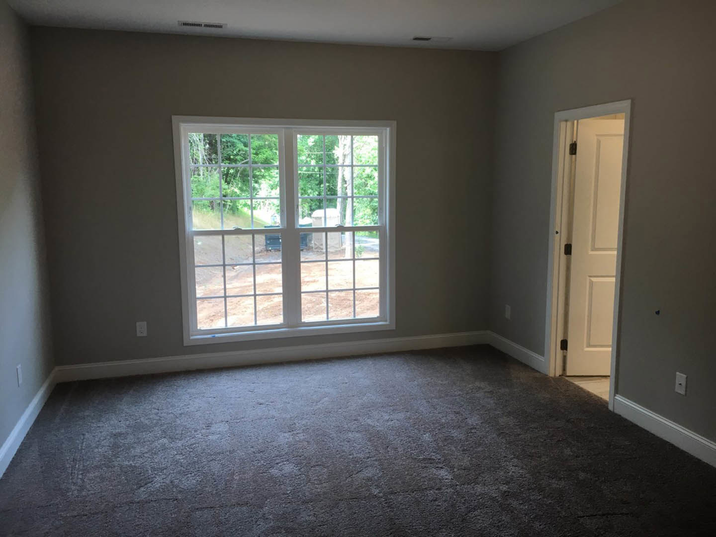 Carpeted bedroom with multi-pane window overlooking trees and dirt, white walls, and open white door with black hinges