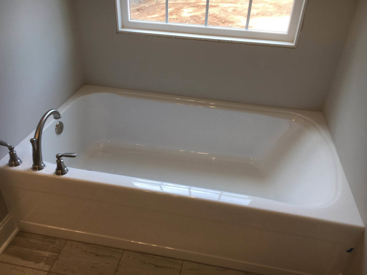 Freestanding white bathtub with chrome faucet beside a window, set on light tile flooring in a modern bathroom