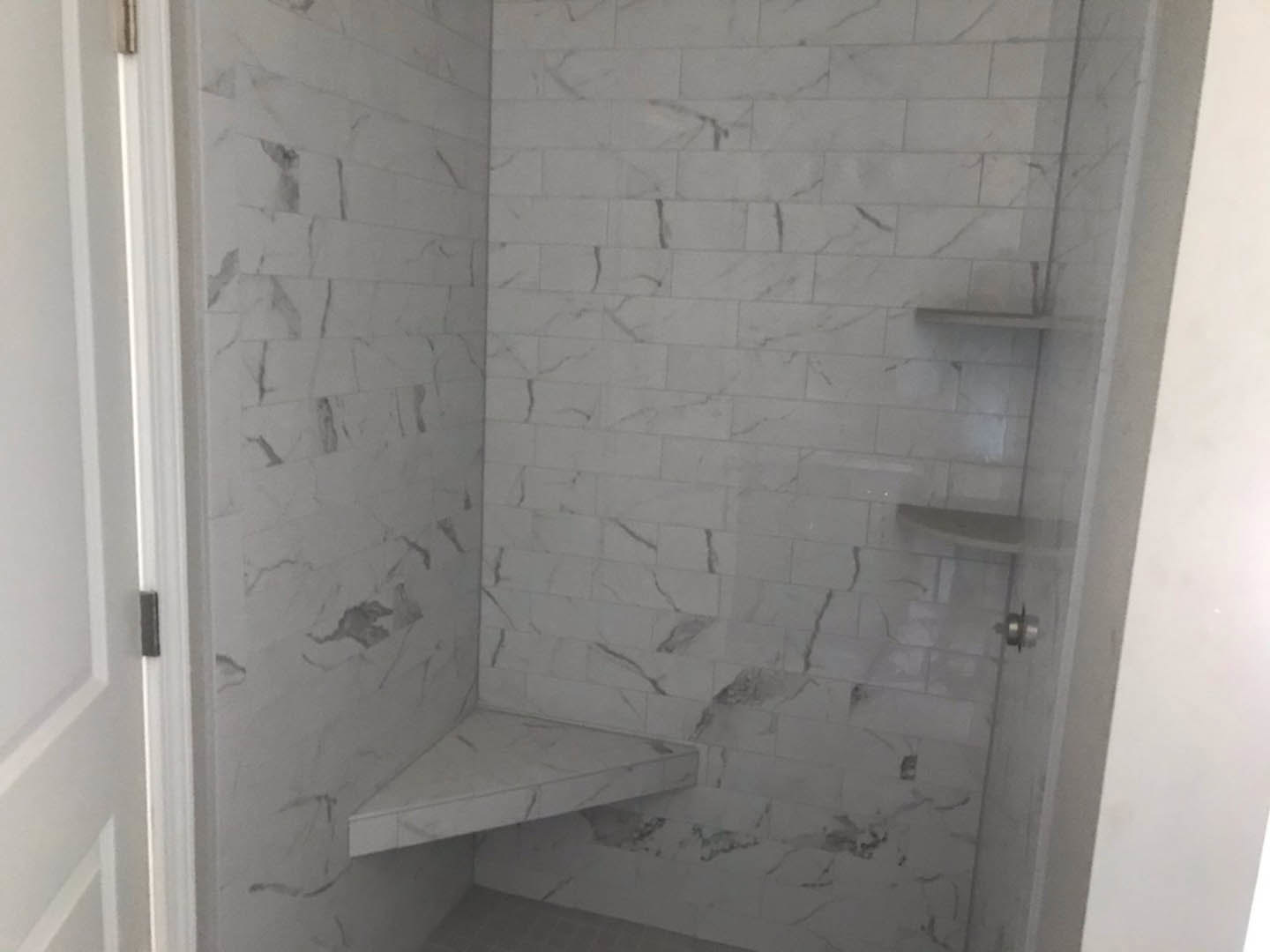 Marble shower wall with built-in shelf, white tile floor, and partial view of white door frame