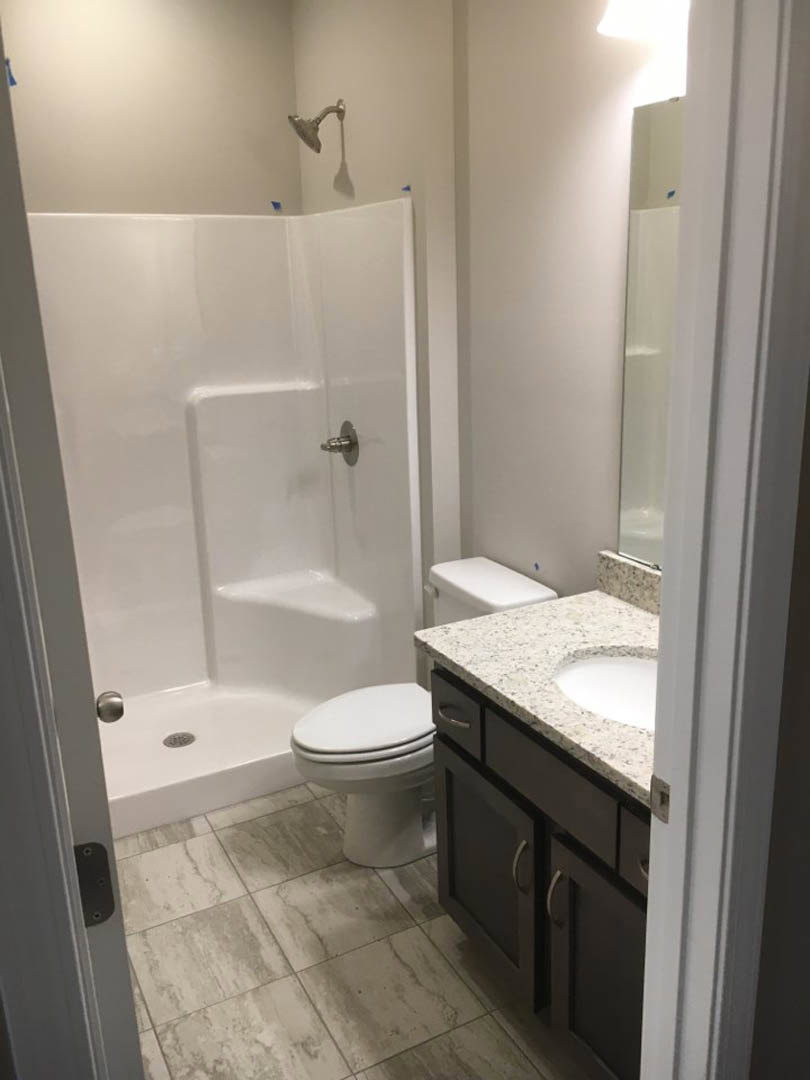 Bathroom with white tile floor, wall-mounted shower head, white toilet beside rectangular sink, large mirror above sink, and white tiled shower enclosure.