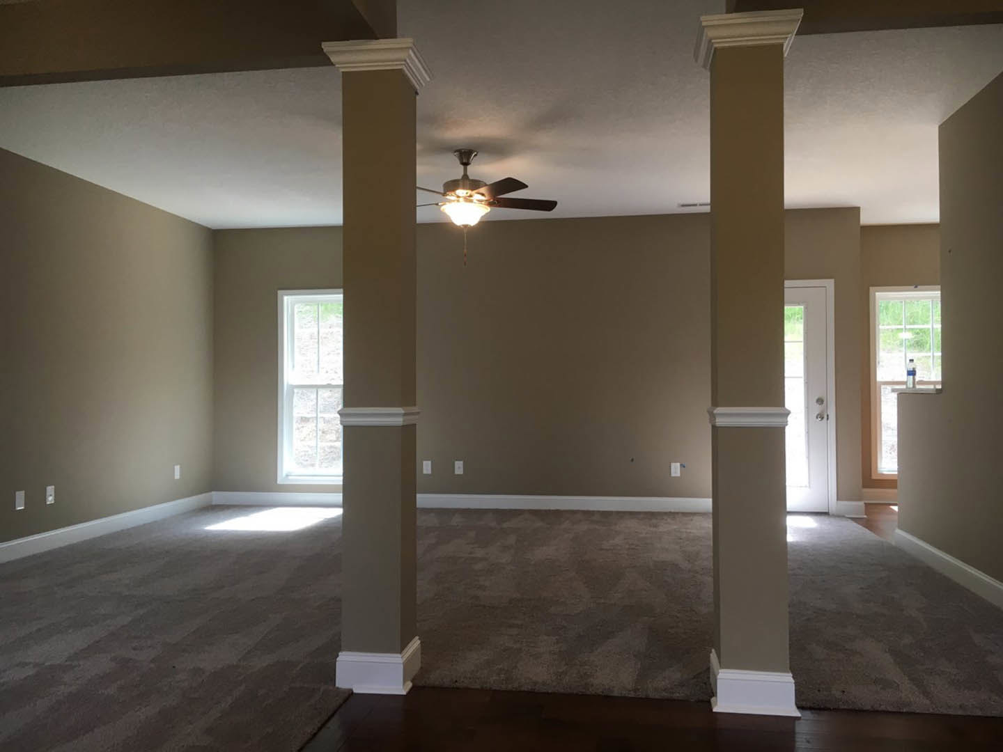 Open living space with white columns, beige carpet flooring, white-framed window, paneled door, ceiling fan with light, and neutral painted walls with crown molding.