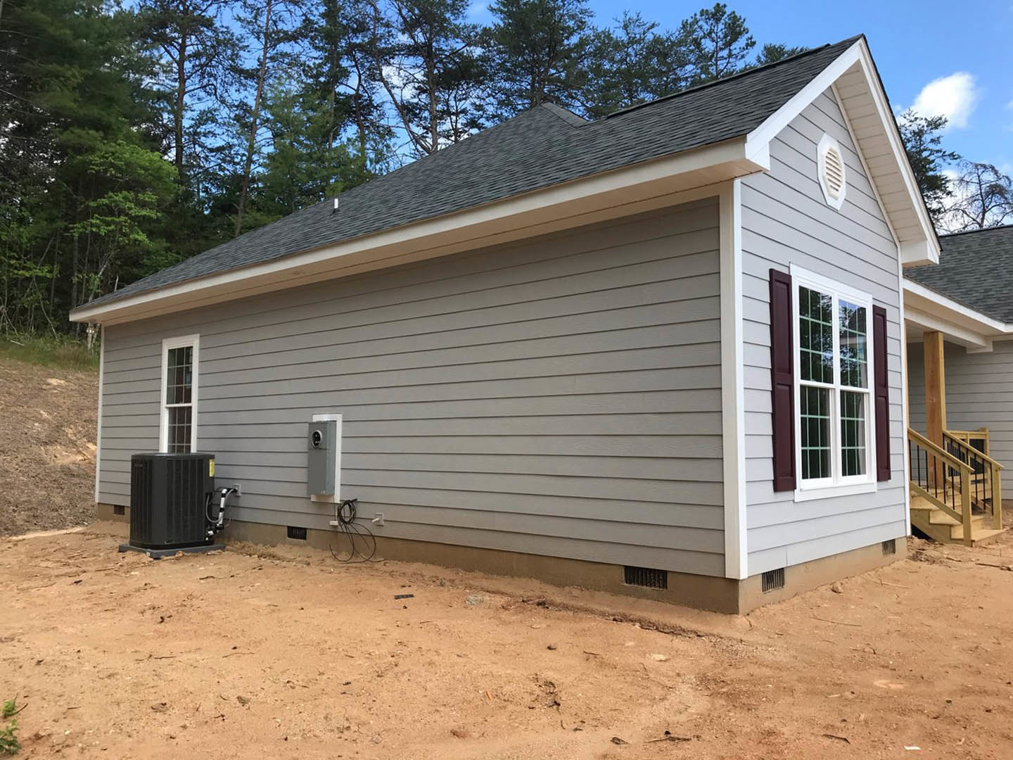 Grey siding exterior with large picture window, white vent, black rectangular utility box with pipe, and wooden staircase featuring metal railings; tree and shed visible in