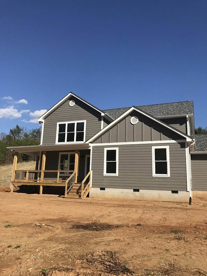Partially built house with white framed windows, wooden porch and staircase, unfinished siding, dirt road in foreground, trees and cloudy sky in background