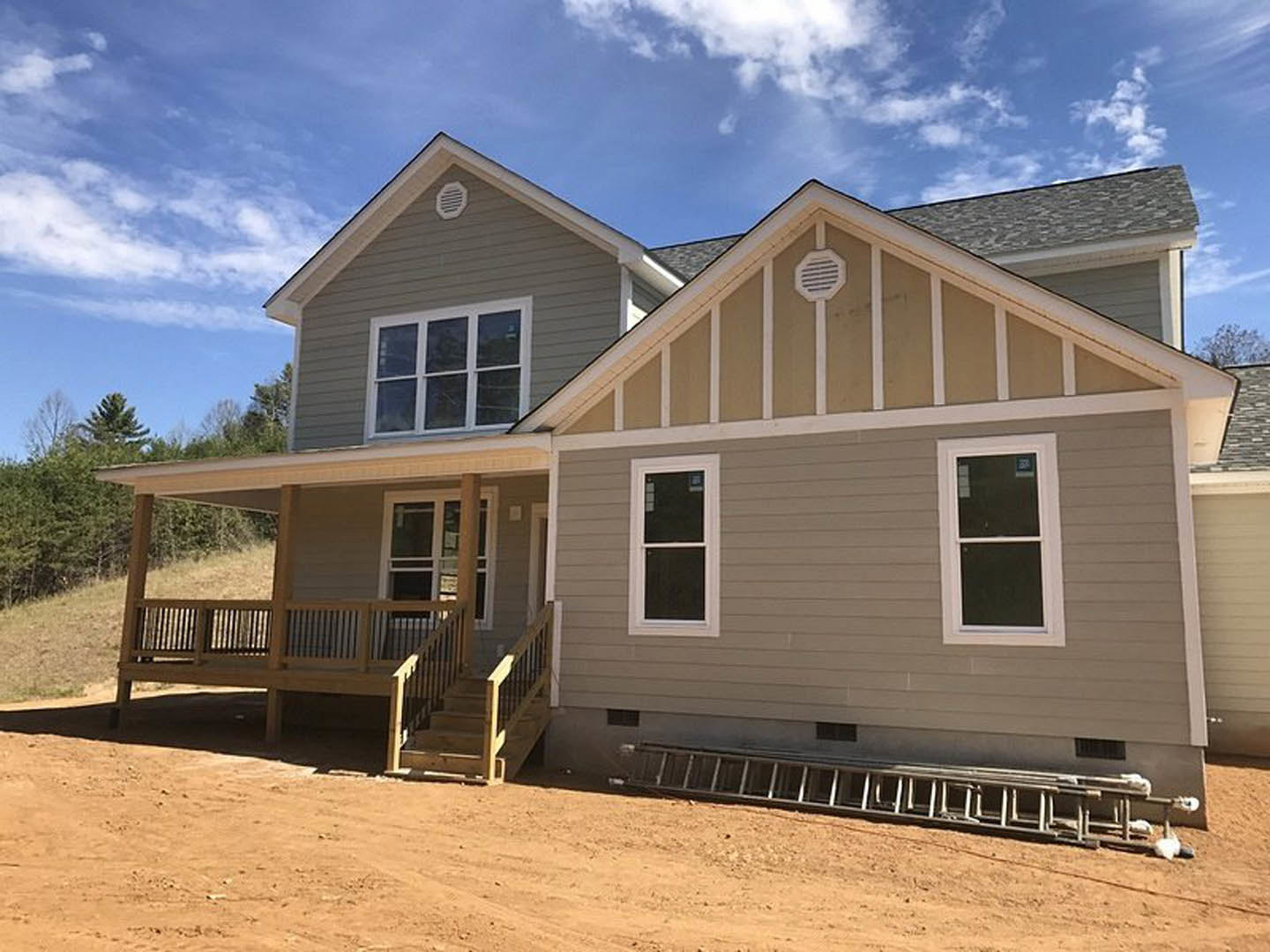 Two-story house under construction with exposed framing, white window frames, exterior siding, dirt road in foreground, blue sky and clouds overhead, ladder leaning against porch