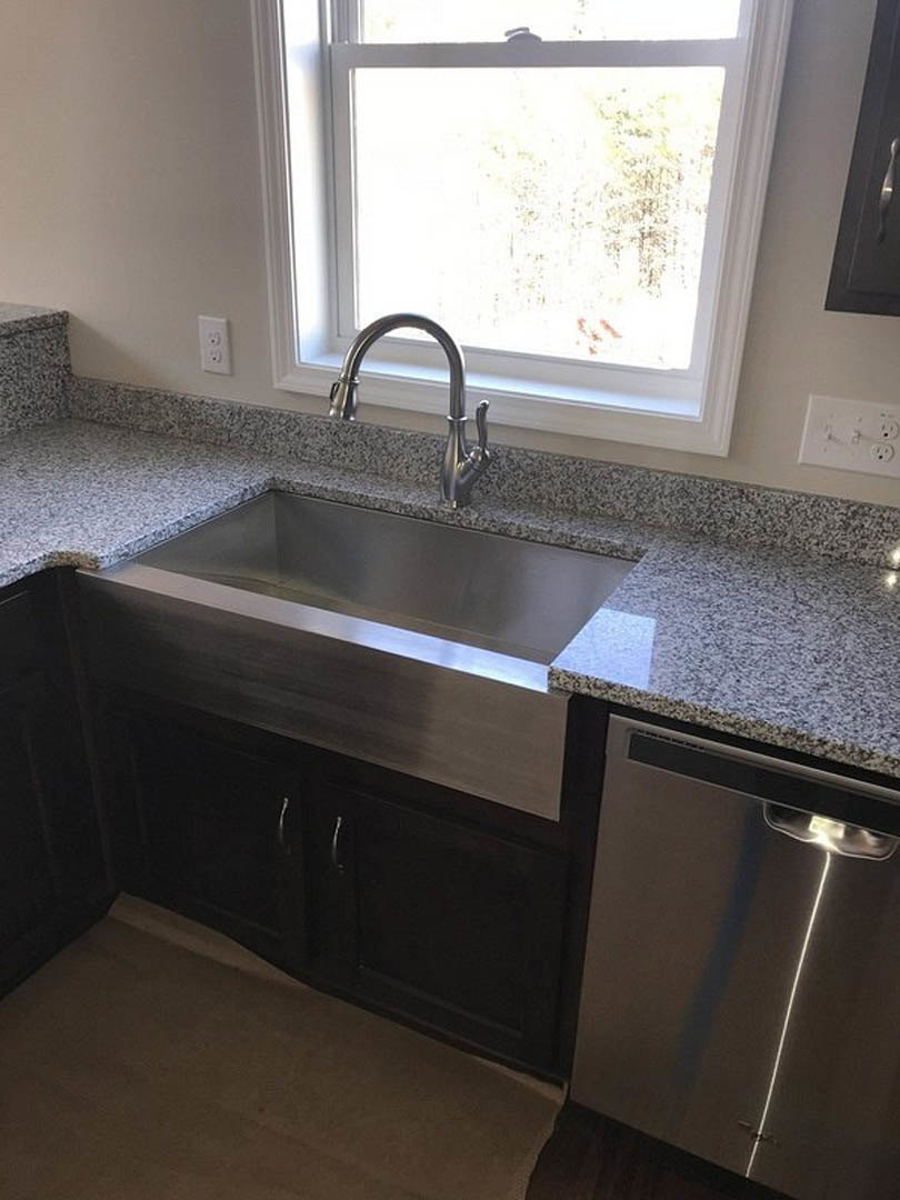 Granite countertop kitchen with stainless steel sink and faucet beneath a window, silver dishwasher, white tile backsplash, and white cabinetry