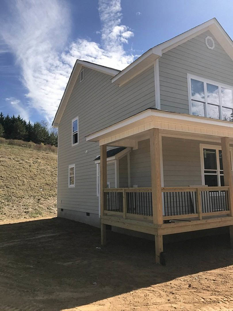 Two-story home with light siding, covered front porch featuring wooden decking and metal railings, white picket fence, large windows, manicured lawn, and trees in the background