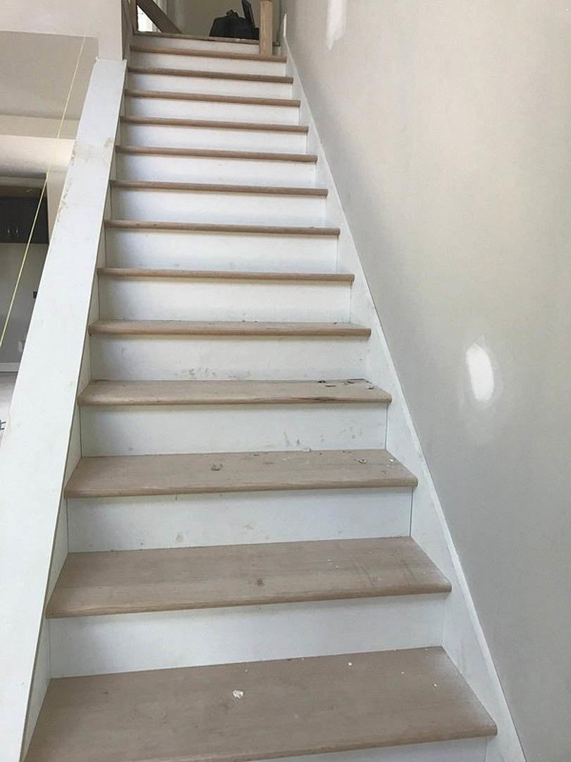 White staircase with natural wood steps, white balusters and handrail, adjacent to smooth gray flooring and white walls.