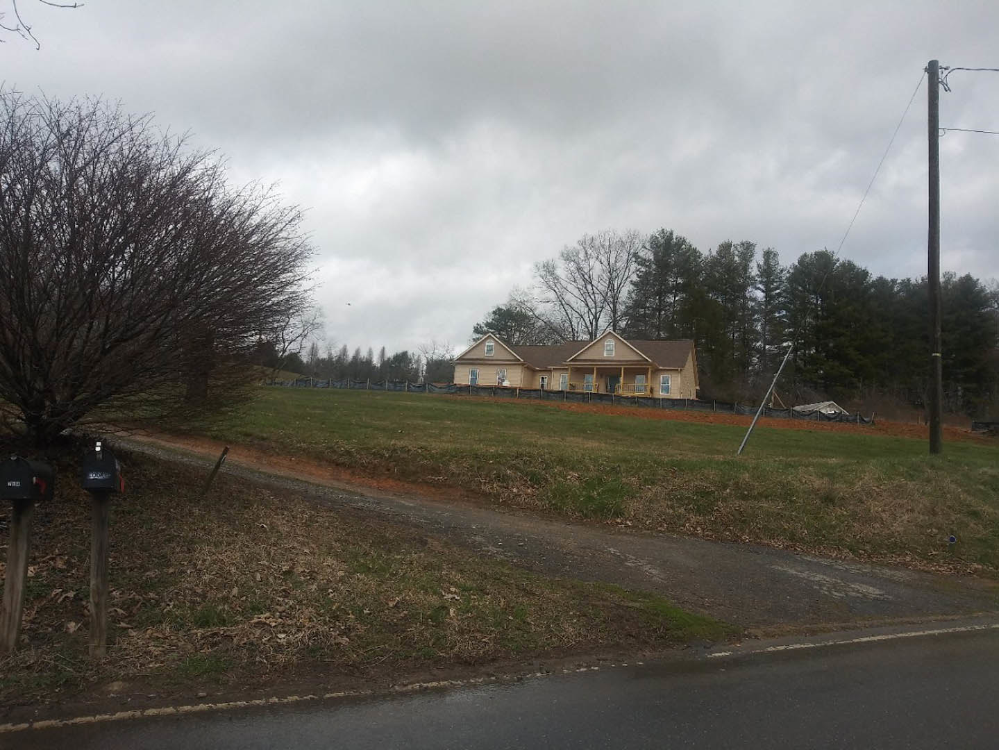 Modern two-story home with light siding and dark roof sits atop a grassy hill, bordered by a wooden fence and surrounded by mature trees; asphalt driveway curves through the