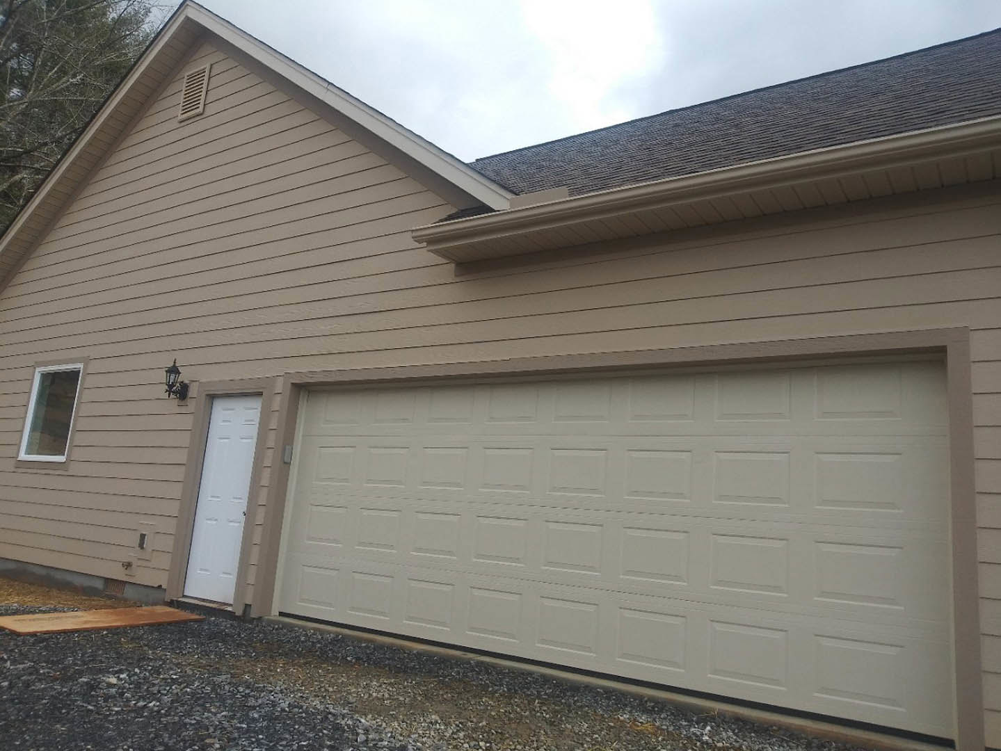 Two-story home with gray siding, brown trim, white front door, attached garage with white door, gravel driveway, large windows, and vent detail near the roof.