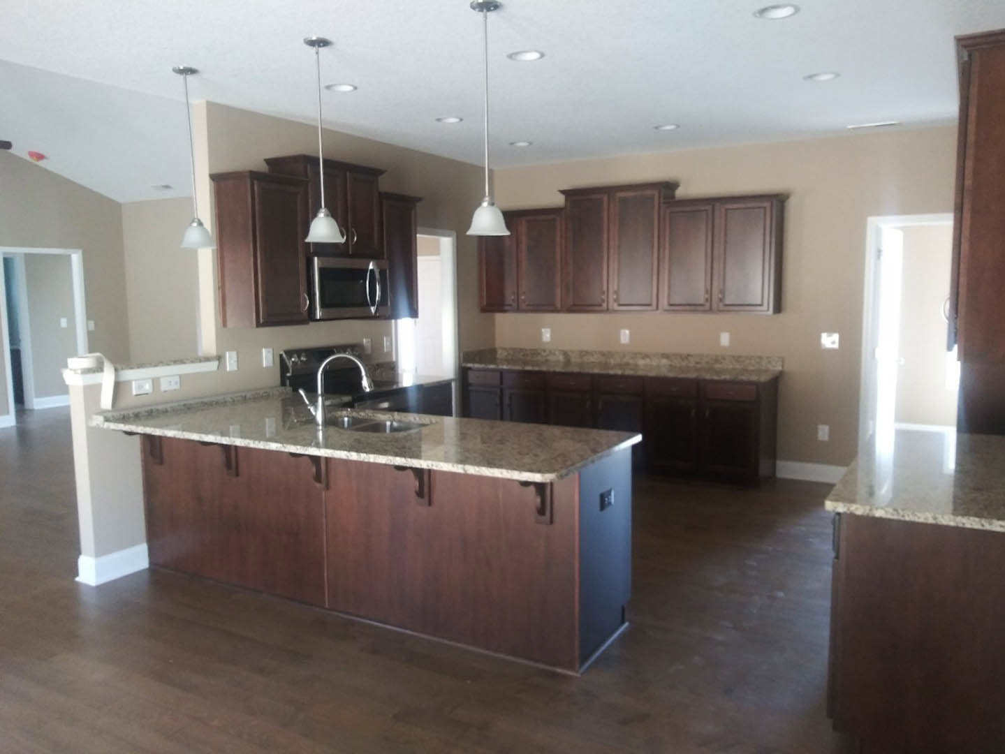 Granite countertops, wood cabinetry, kitchen island with built-in sink, stainless steel microwave, tile flooring, white wall, ceiling lighting