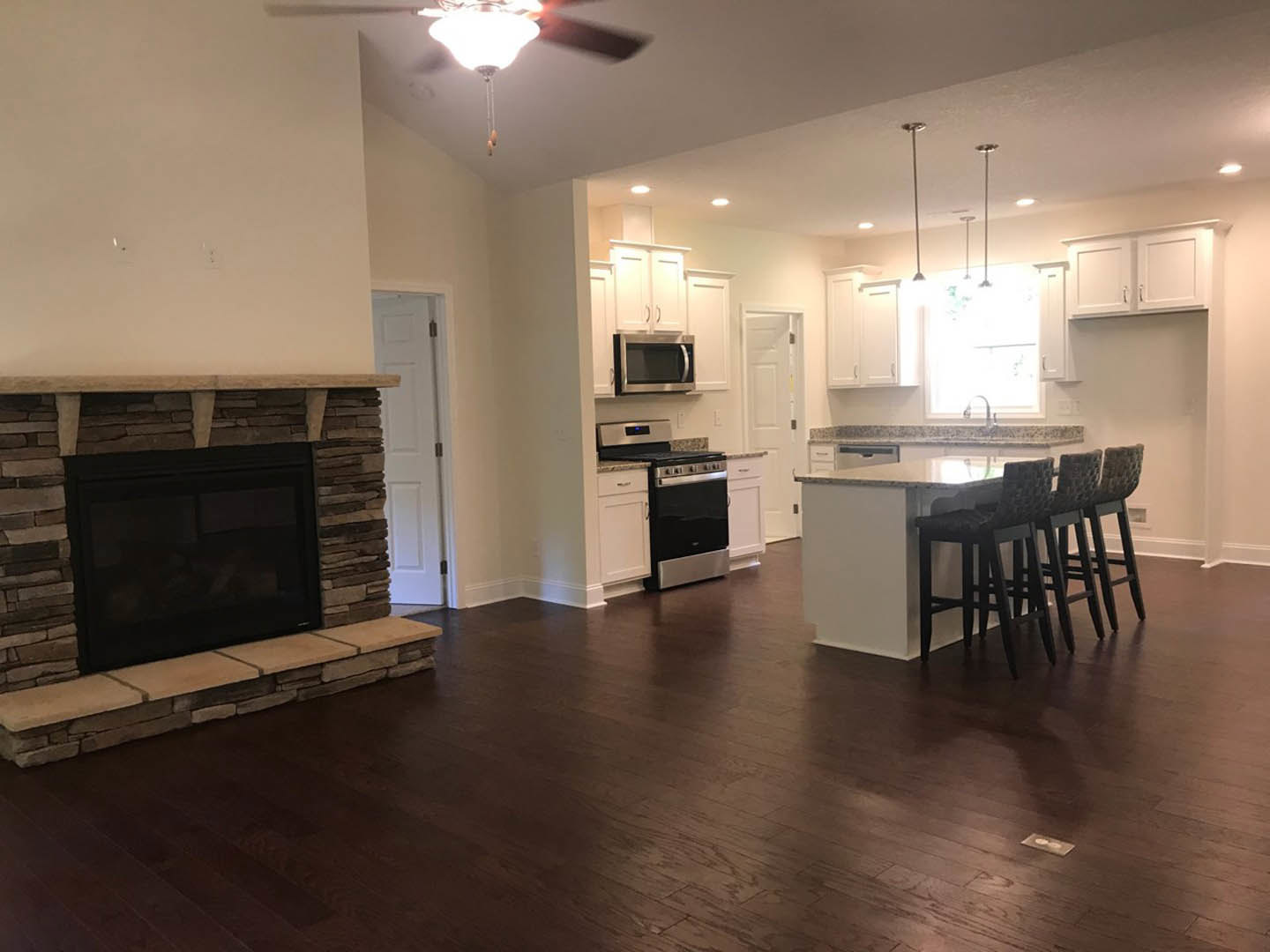 Open-concept kitchen and living room featuring stone fireplace, hardwood floors, bar stools at island, white cabinetry, stainless steel appliances, and open microwave door