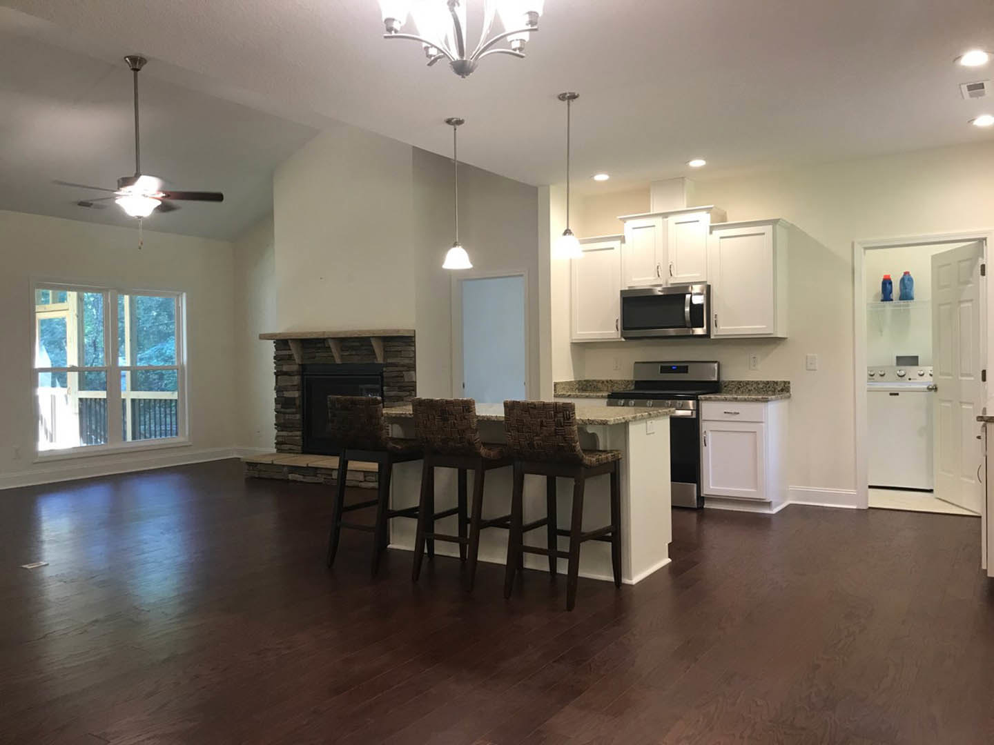 Open-concept kitchen and living room featuring a stone fireplace, white cabinetry, stainless steel microwave, bar stools at a quartz island, wood flooring, and modern light
