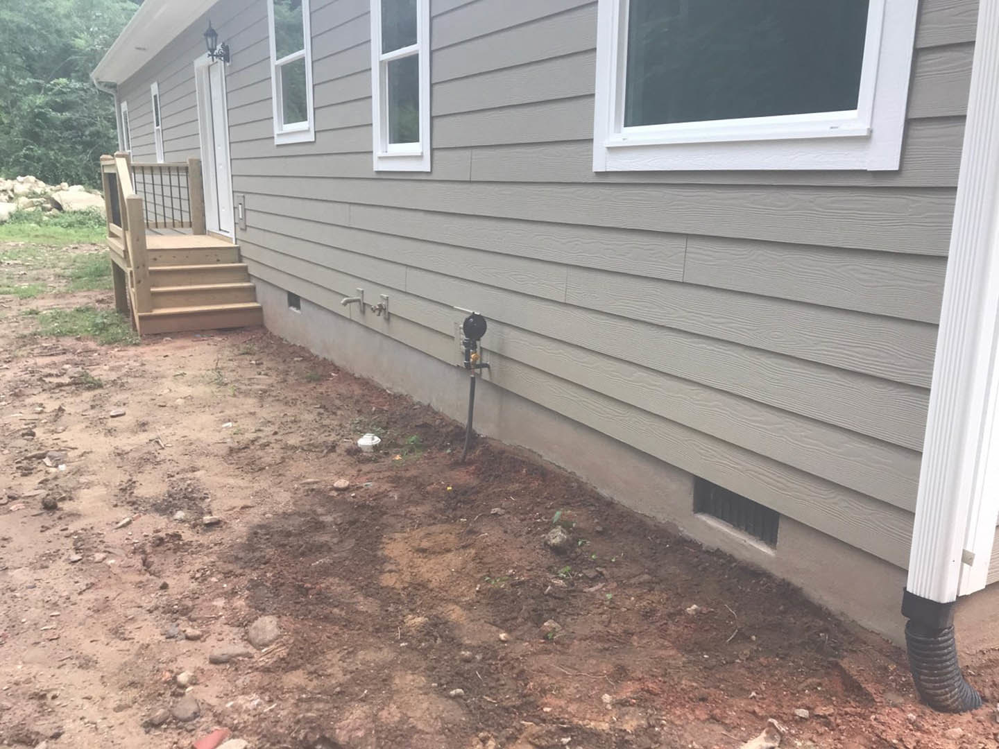 Grey siding custom home with white-framed windows, wooden deck and porch, exterior wooden stairs, black pipe with yellow handle, backyard landscaping.