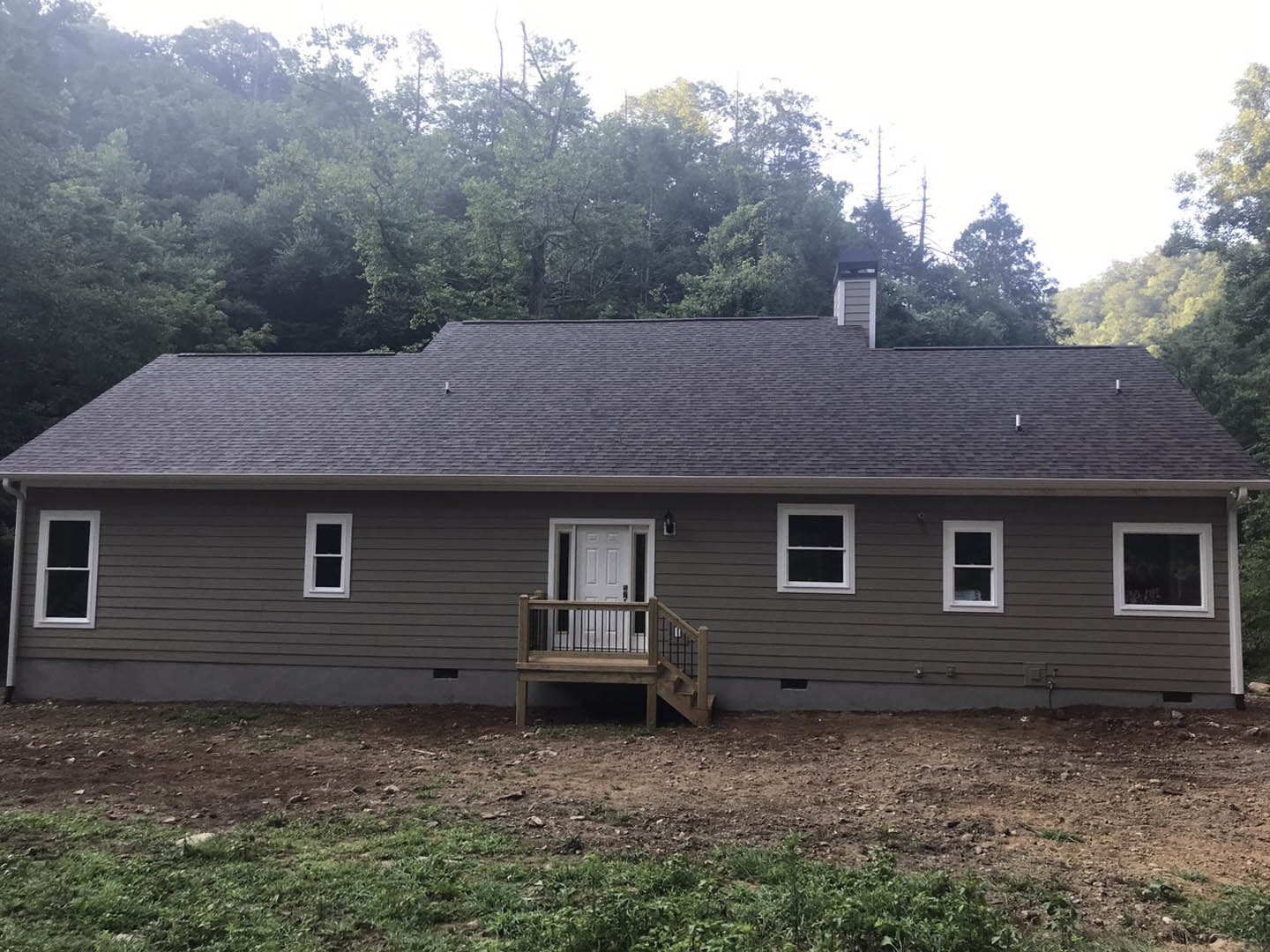 White siding house with a covered porch, wood deck, white framed window, wooden bench near the front door, wooden stairs leading to a grassy yard, and shingled roof.