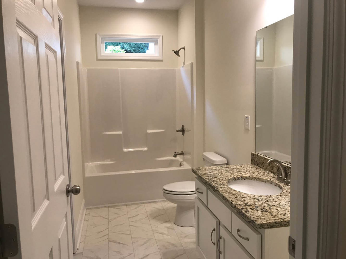 Modern bathroom with white porcelain sink and toilet, light gray tile walls, chrome faucet, window overlooking green trees, glass shower enclosure, and brushed metal door knob