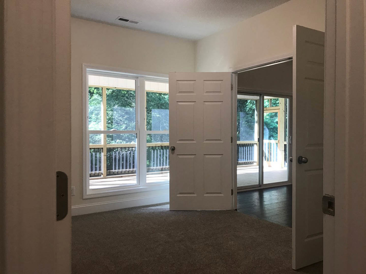 White paneled door with black handle open to carpeted room, window with metal railing and leafy trees visible outside, deck beyond doorway.