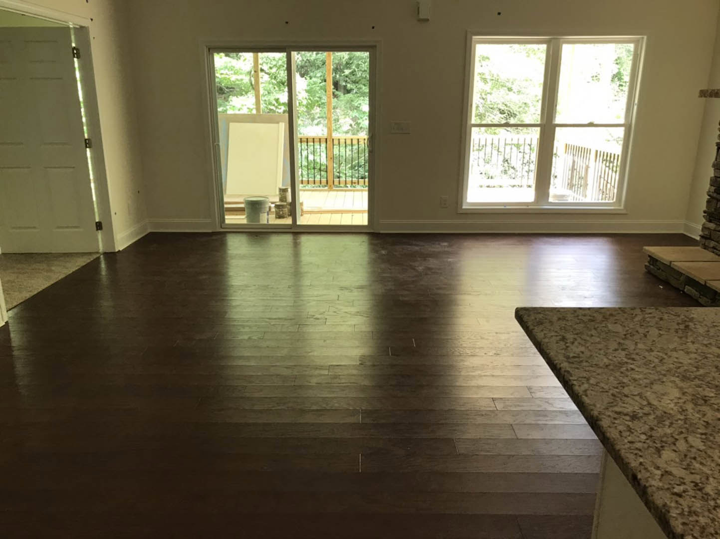 Marble countertops with white cabinetry, hardwood flooring, sliding glass door opening to deck, large window with railing and trees visible outside, glass and cup on counter
