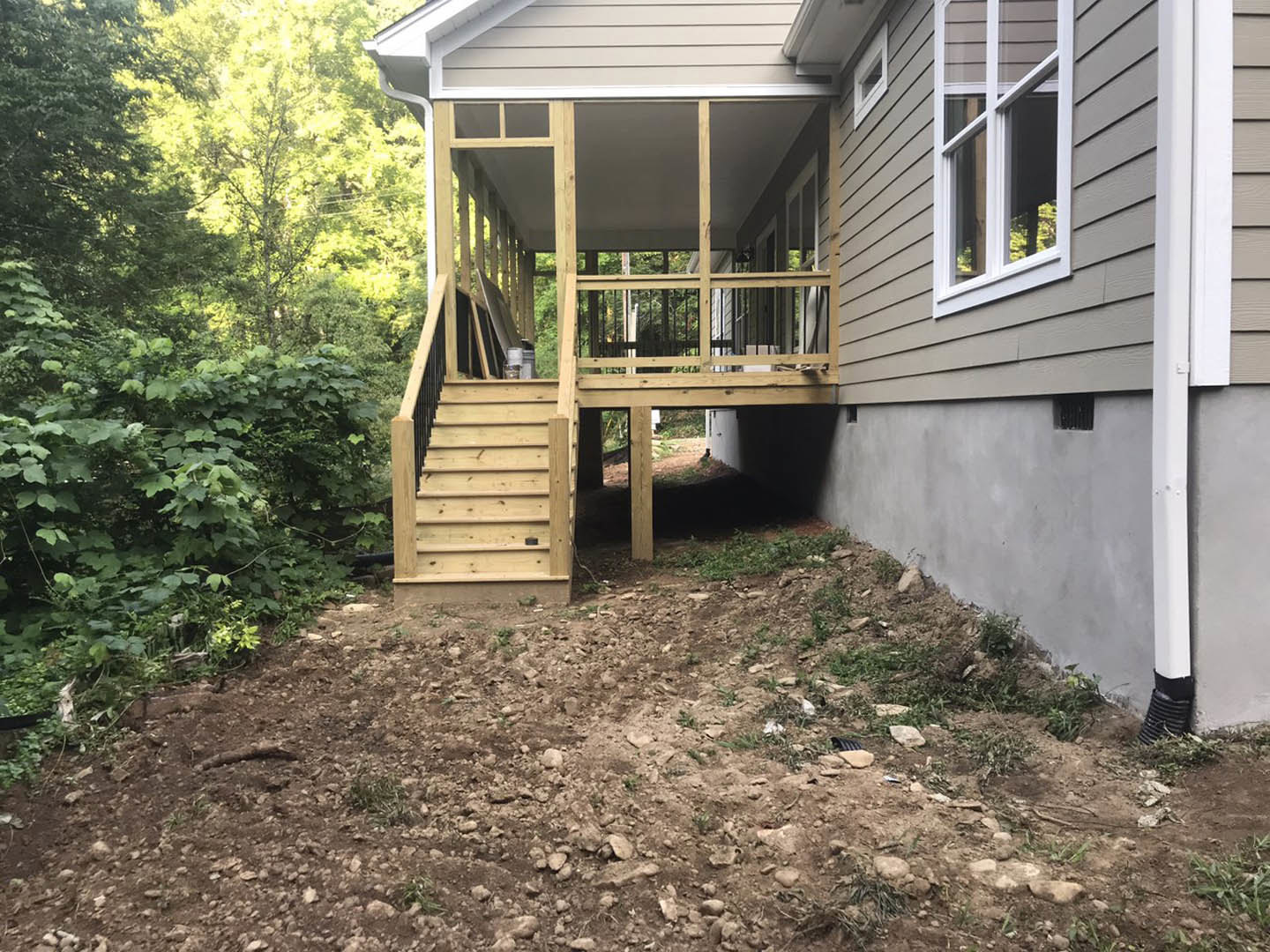 Two-story home with wood deck and stairs, white-framed windows, metal drain, dirt yard, surrounded by tall trees.