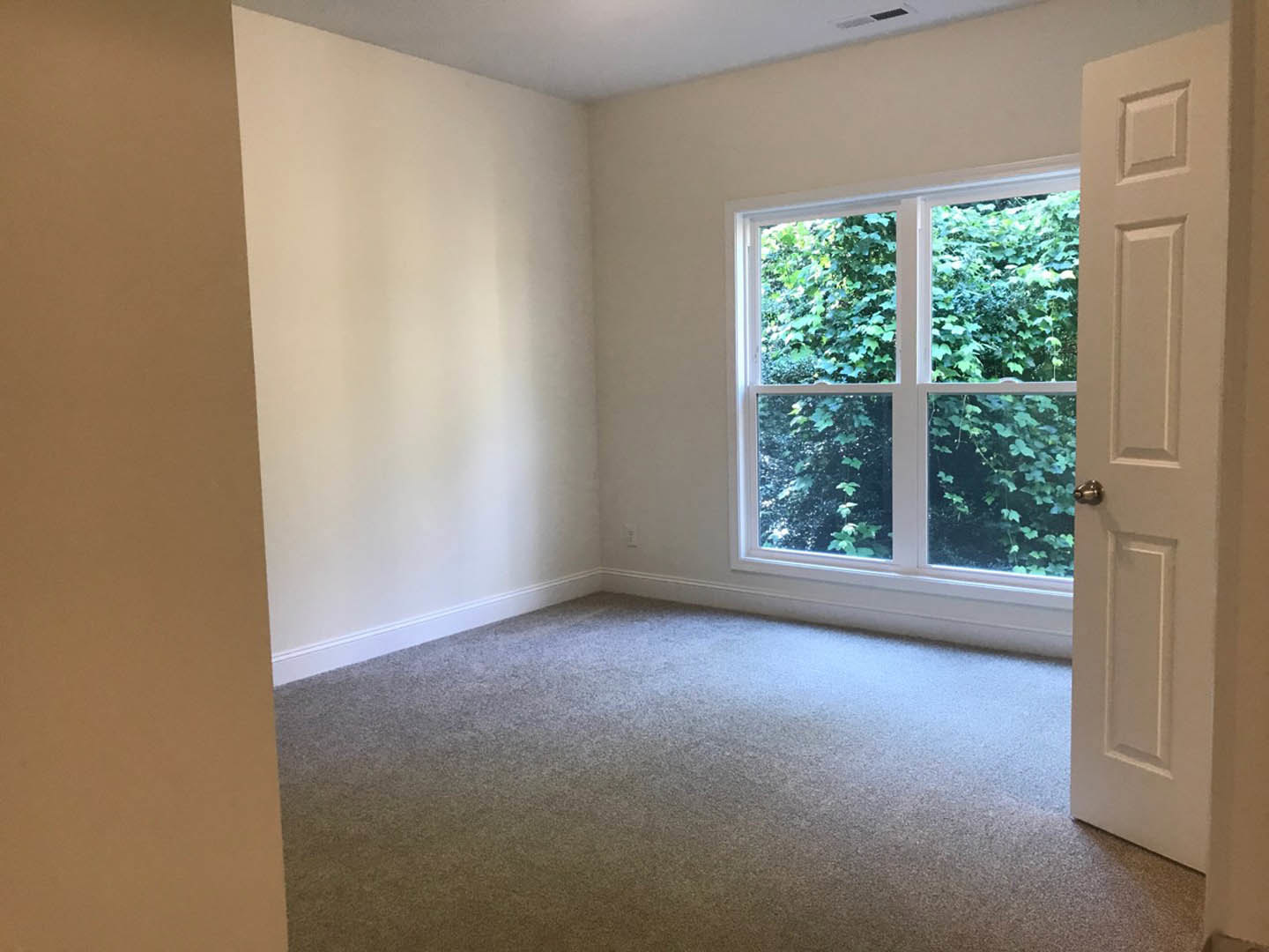 Carpeted bedroom with open white door, large window overlooking green trees, neutral walls, and wood trim