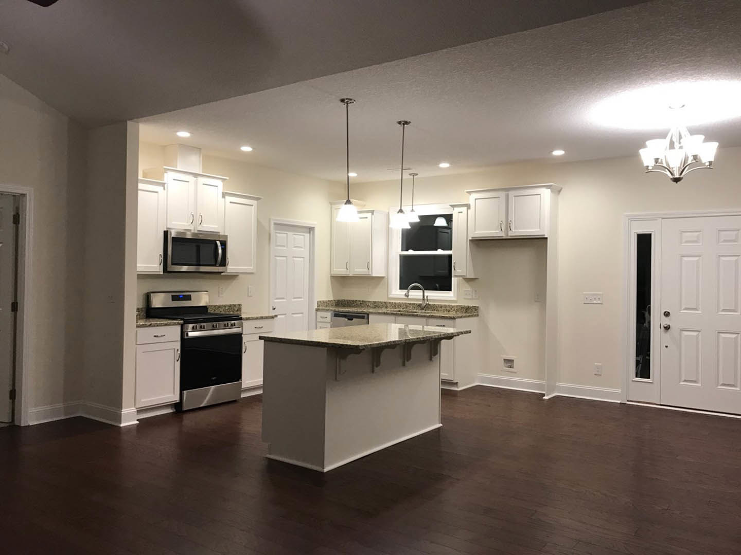 White kitchen with granite countertops, dark wood flooring, built-in microwave, stove and oven, bar seating, white cabinetry, and a white door with a doorknob.
