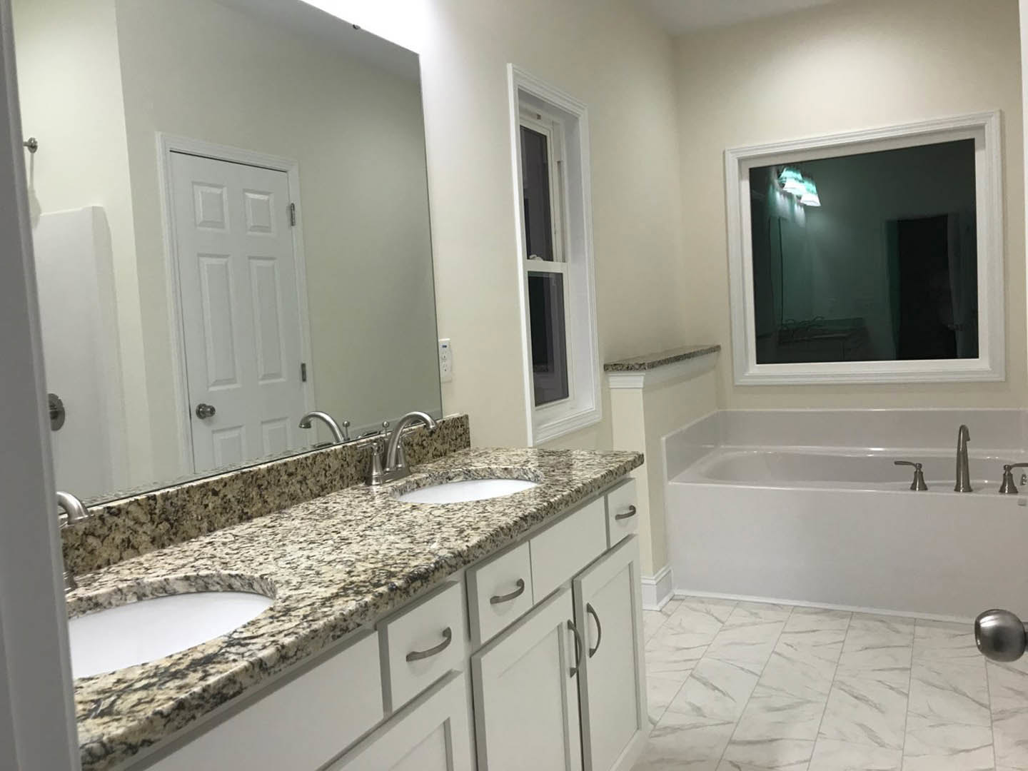 Bathroom featuring marble countertops with dual sinks, freestanding bathtub with chrome faucet, white door with metal handle, large window allowing natural light, and tiled walls.