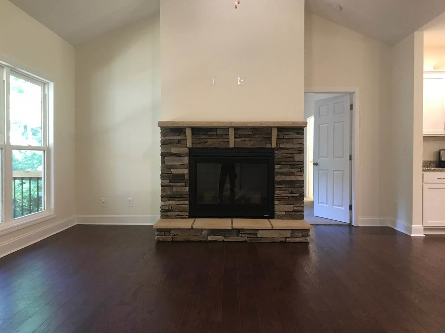 Modern living room with wood flooring, white-framed window, glass door reflecting a person, central fireplace with stone hearth, and white door with silver doorknob