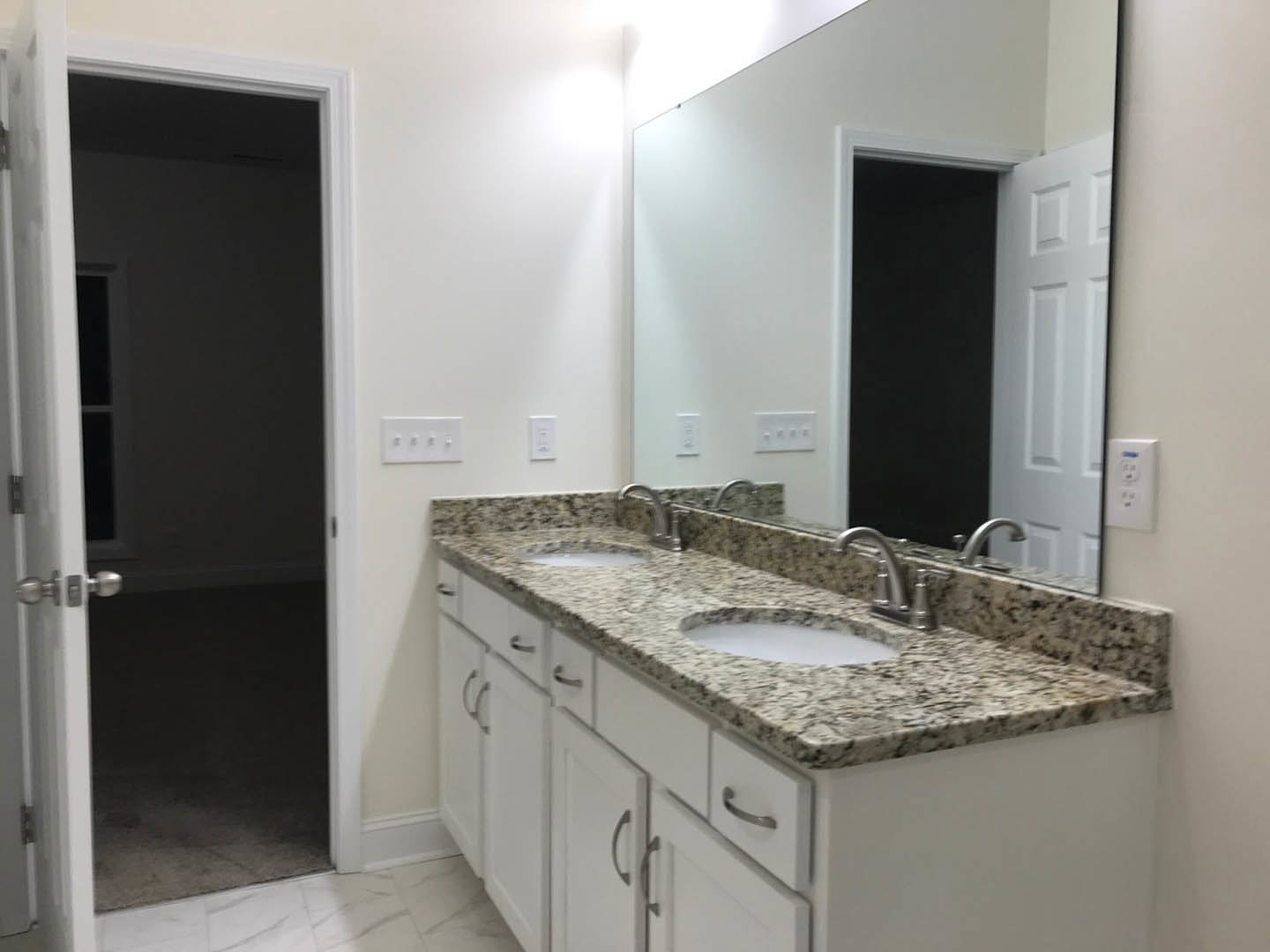 Marble bathroom countertop with undermount sink, chrome faucet, large wall mirror, white cabinetry, and tile backsplash; open door leads to a dimly lit adjacent room.