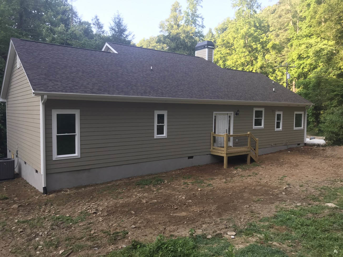 Two-story house with gray siding, white-trimmed windows, covered front porch with wooden railing, brick chimney, landscaped yard with grass and dirt patch, mature trees in