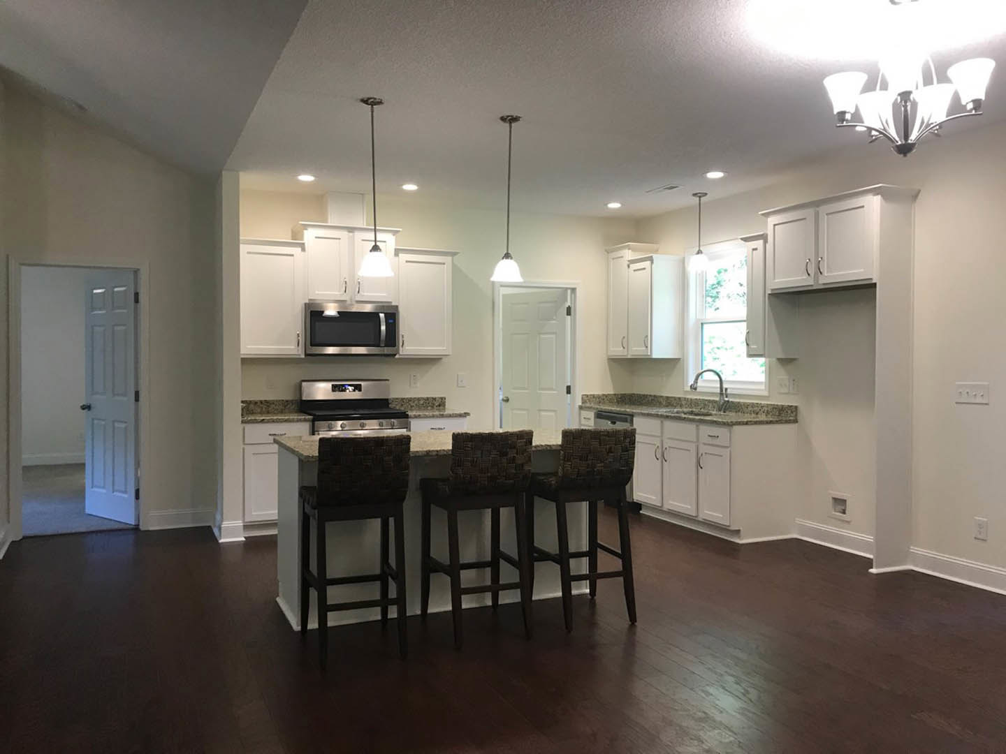Open-concept kitchen featuring wood flooring, a row of black bar stools at a white quartz island, shaker cabinetry, stainless steel appliances, and recessed lighting