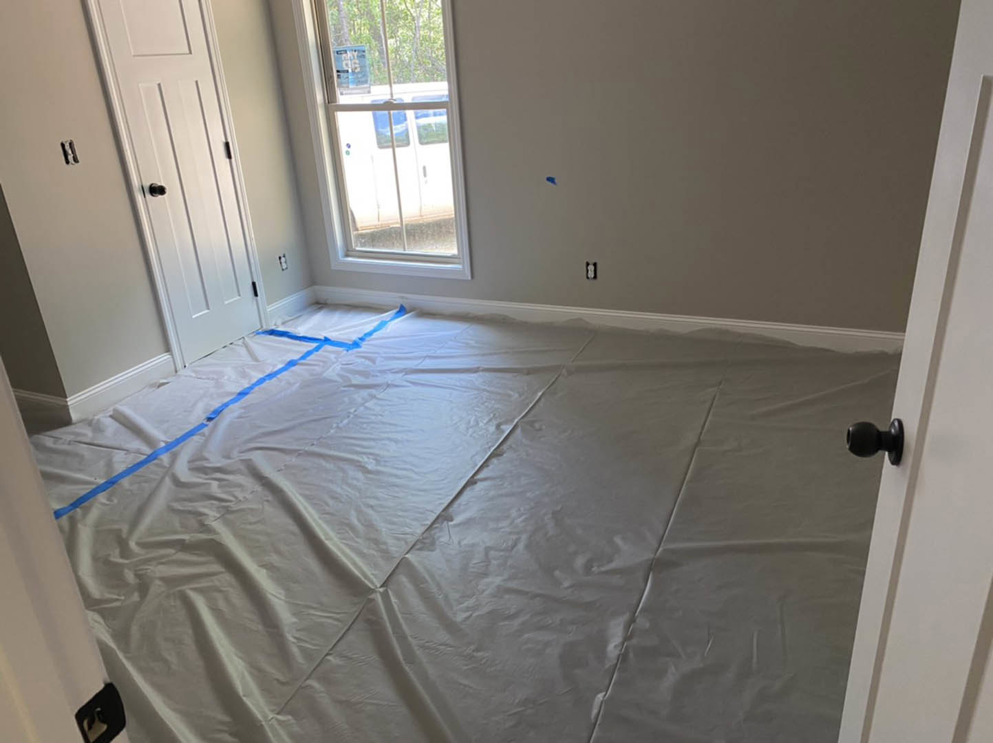 Unfinished room with white sheet taped to floor, white door with black knob, window, and light-colored plaster walls