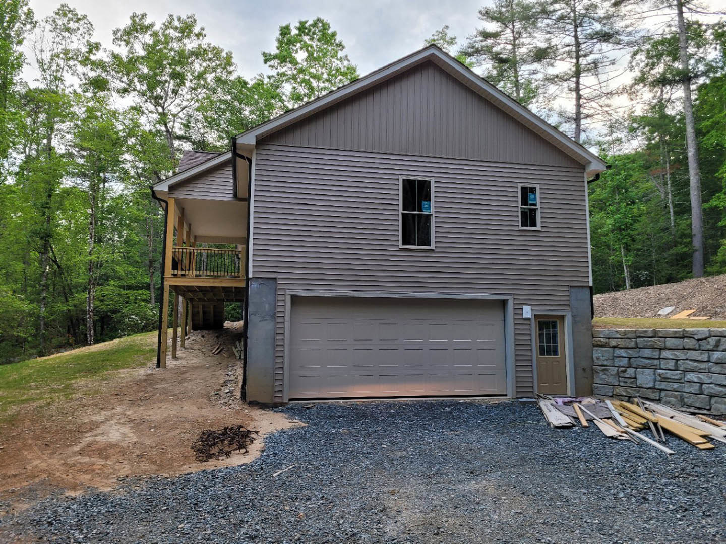 Two-story house with attached garage, concrete driveway, white garage door, front window displaying a blue sign, scattered wood pieces on ground, surrounded by trees and lawn.