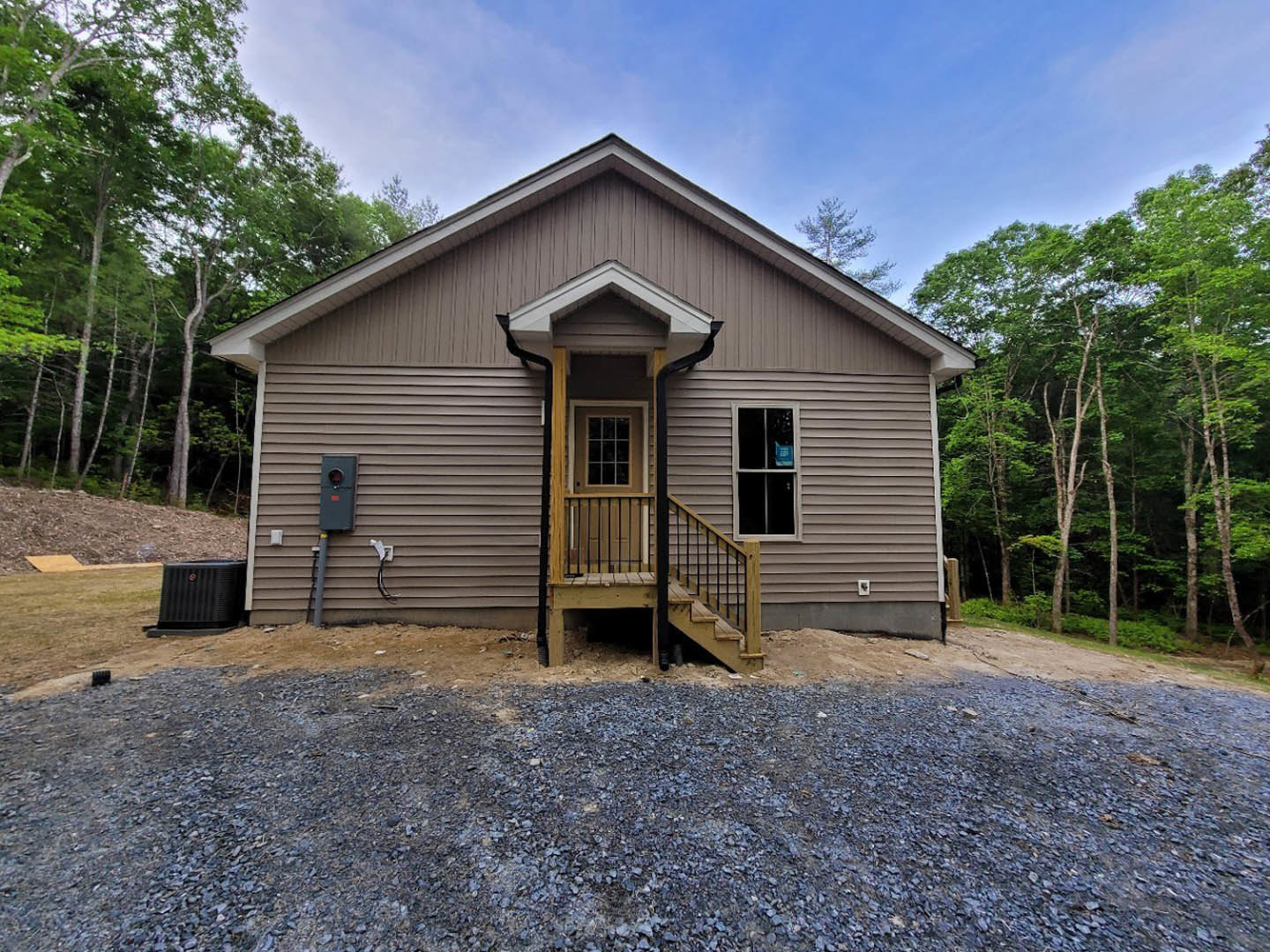 Two-story cottage with wood siding, gravel driveway, covered front porch with stairs, surrounded by mature trees and landscaping rocks.