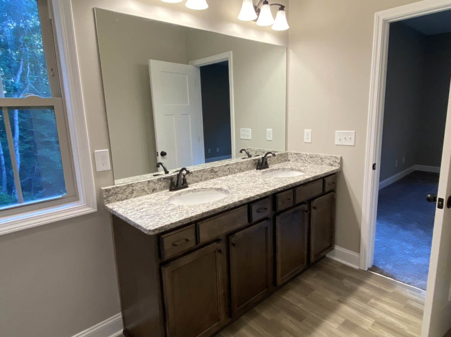 Bathroom with double vanity featuring undermount sinks, wide mirror above, white outlet and three-knob light switch on tiled wall, window with white frame, white door with black