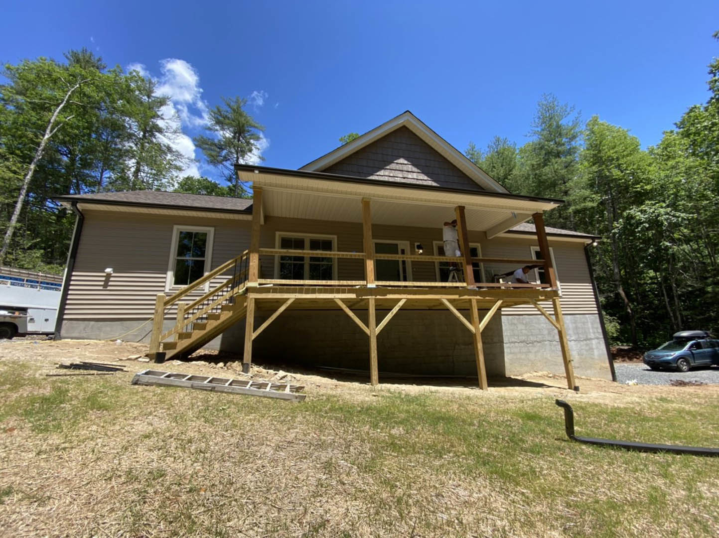 Two-story house with covered front porch, wooden deck, and large windows reflecting trees; man standing on deck, car with roof rack parked nearby, grassy yard with wooden ladder