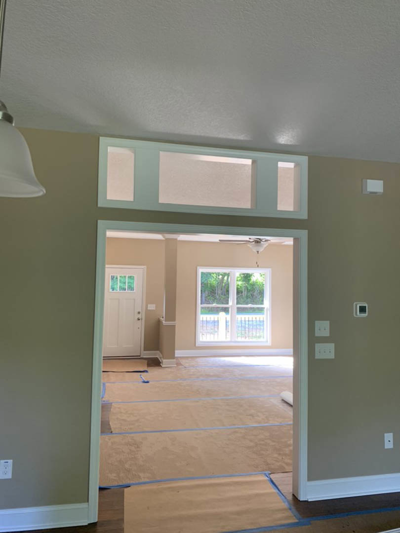 White door with glass window opening into a room with light-colored plaster walls, white-framed window, and carpeted floor marked with blue tape.