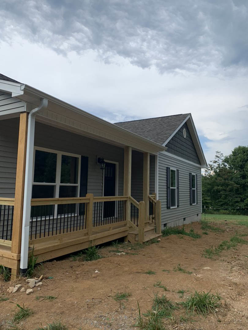 Two-story home with light siding, covered front porch featuring wooden railing, fenced yard, leafy tree, stone landscaping, and cloudy sky overhead