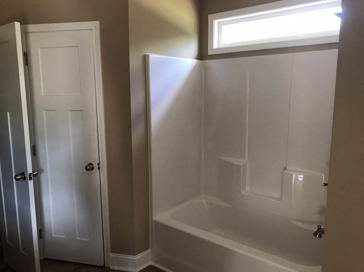 White bathtub with chrome faucet beside a white paneled door with silver handles, wall-mounted light fixture above, neutral tile flooring and soft gray walls.