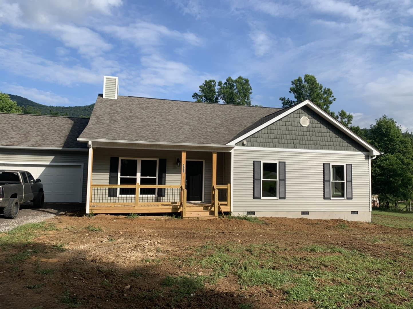 Two-story house with covered front porch, attached garage, gray siding, white trim, large windows, grassy yard, pickup truck parked on driveway, person standing near front door