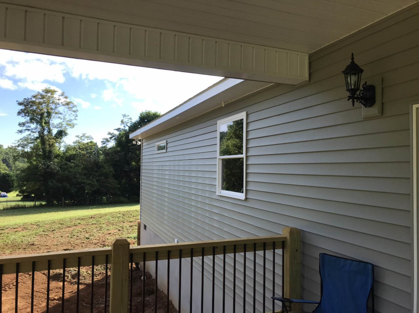 White siding house with covered porch, wooden and metal fence, blue chair with black armrest, wall-mounted light fixture, tree with green leaves visible through window, landscaped