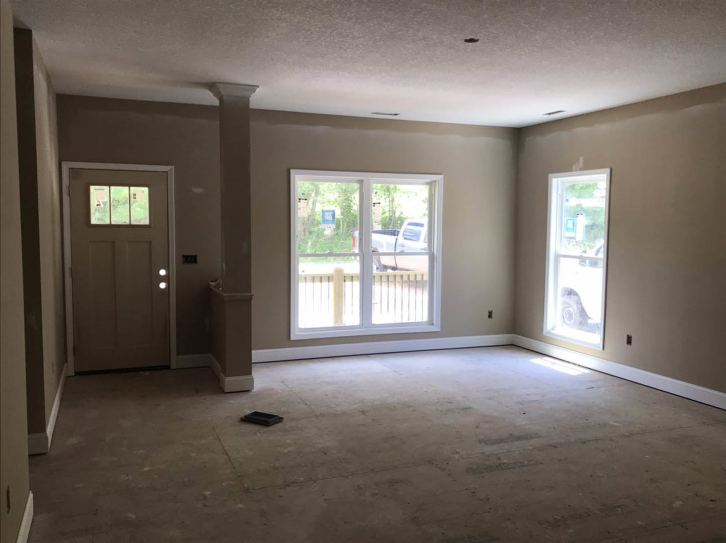 Bright room with white-framed windows, white door with glass panel, light gray walls, smooth ceiling, hardwood flooring, and a vacuum cleaner placed near a black rectangular