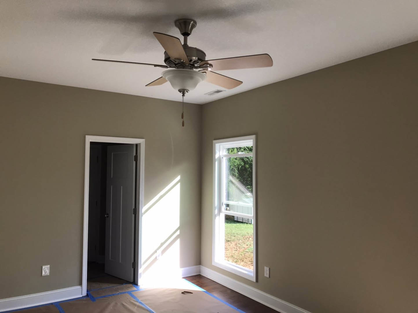 Ceiling fan with integrated light fixture mounted on white plaster ceiling, window with white trim and paneled door visible, neutral walls and baseboard molding, hardwood floor.