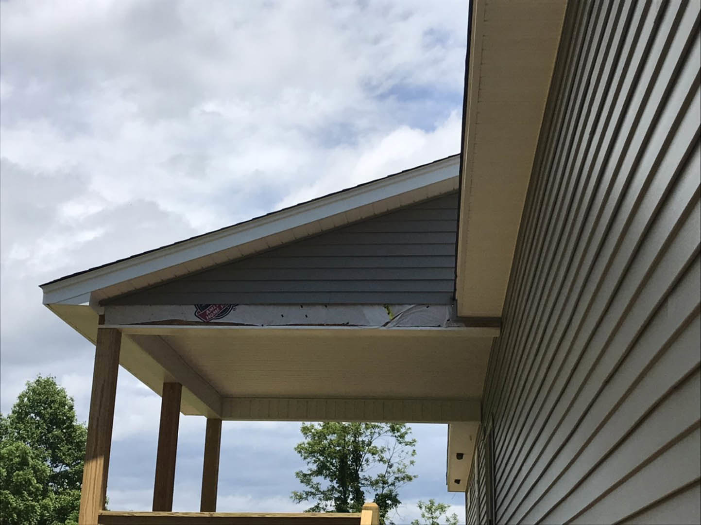 Gabled roof with composite shingles, wide roof overhang, porch area beneath, large window, leafy green tree in front, partly cloudy sky overhead