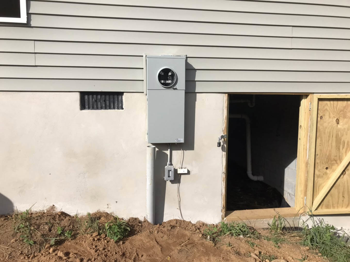Grey electrical box mounted on exterior wall of house above dirt patch, surrounded by plants and wood paneling.