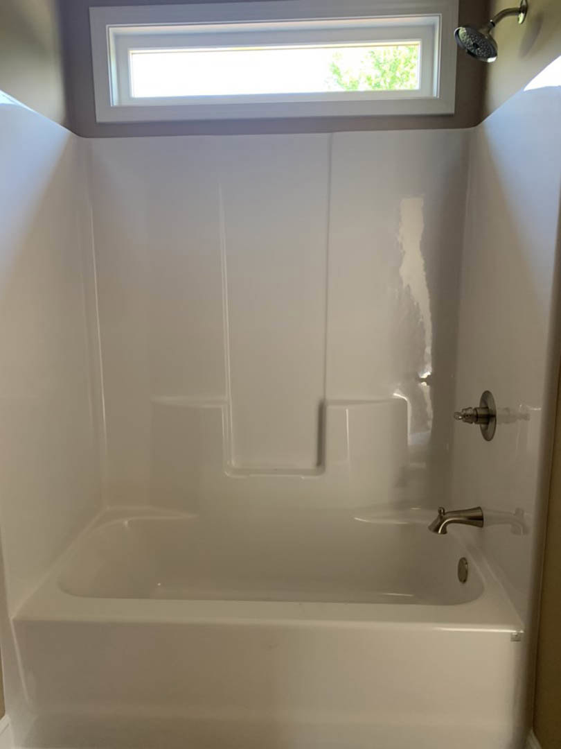Freestanding white bathtub with chrome water spout, light reflecting on smooth tile walls, shower head and green plant visible in modern bathroom