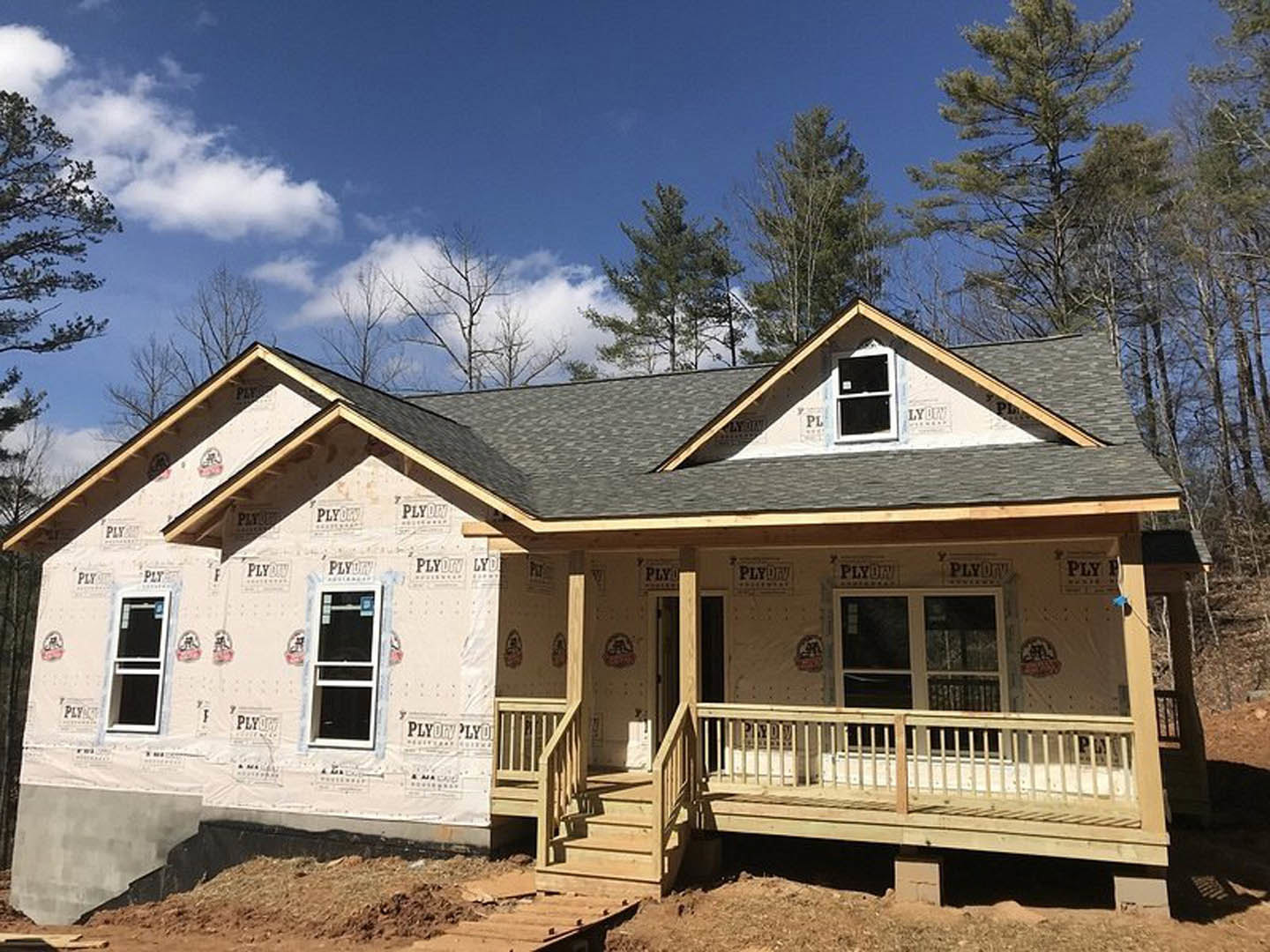 Wood-framed house under construction with covered porch, white door with black screen, white-trimmed window, stairs leading to entry, gray siding, shingled roof, leafy tree in