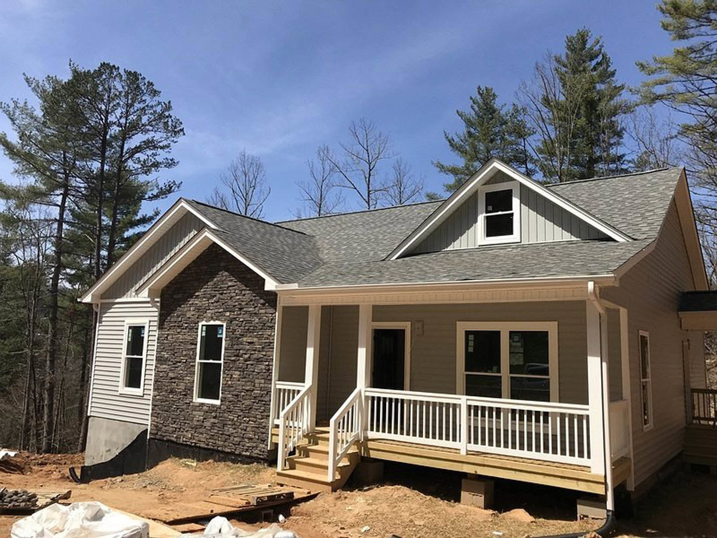 Partially built house featuring a covered porch with white railings, wooden stairs, white-framed windows, and light siding, surrounded by trees under a clear sky.