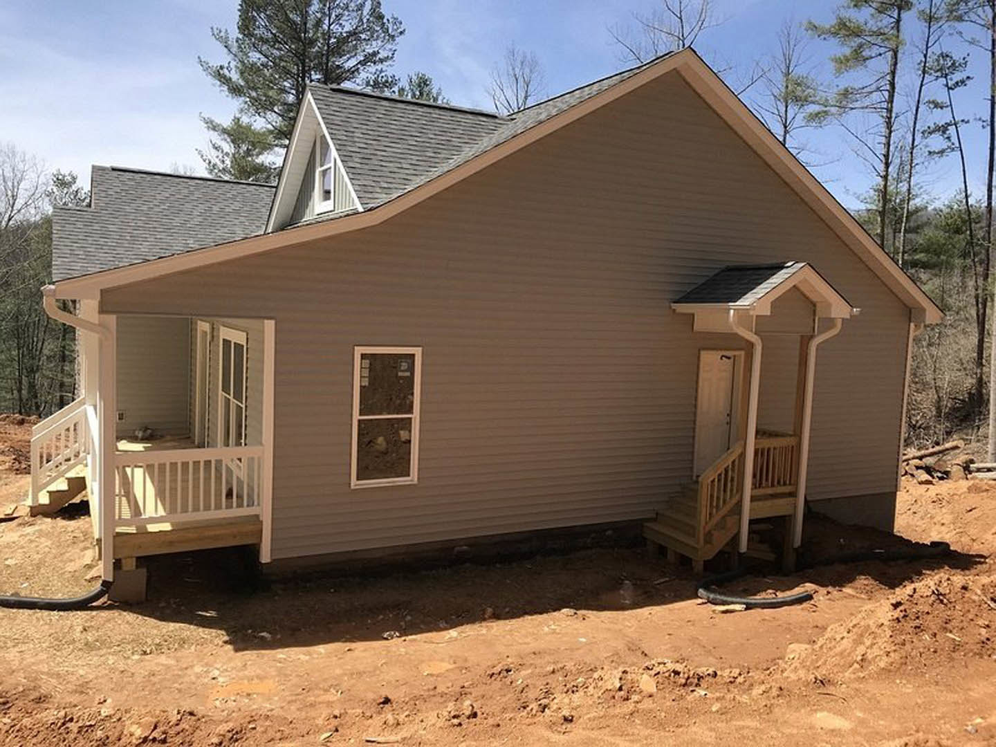 Two-story house under construction featuring a covered porch with wooden railings and stairs, white-framed windows, unfinished siding, and a dirt driveway leading to the property.