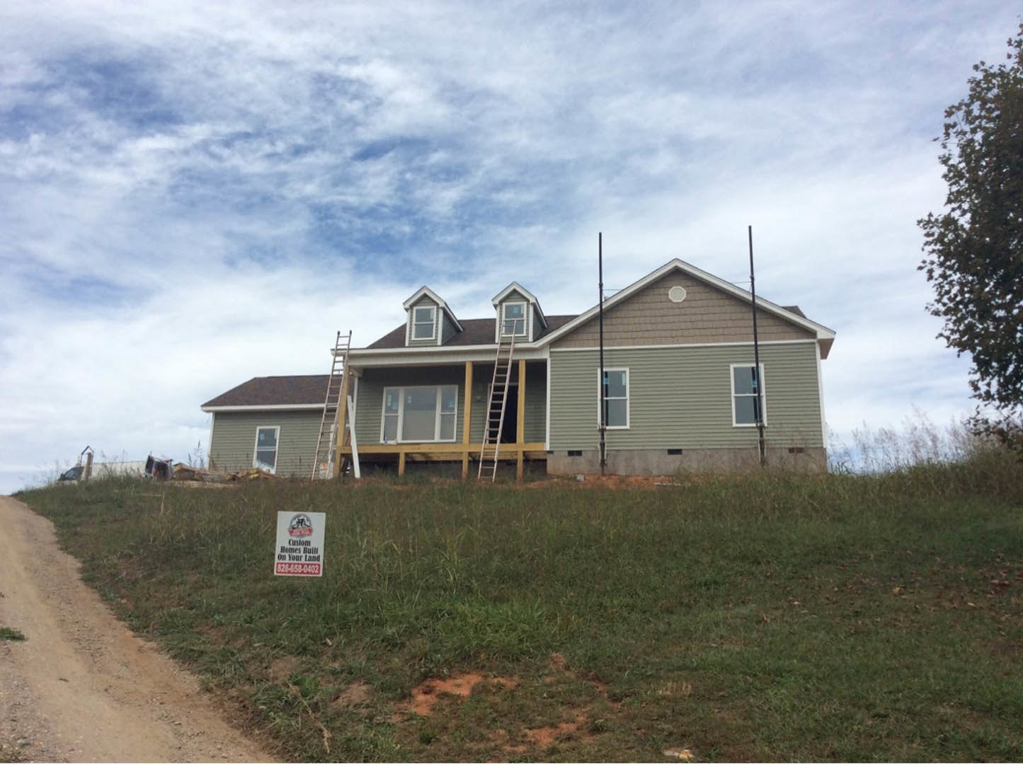 White siding house under construction with multiple ladders leaning against exterior walls, grassy lawn in foreground, windows installed, cloudy sky overhead.