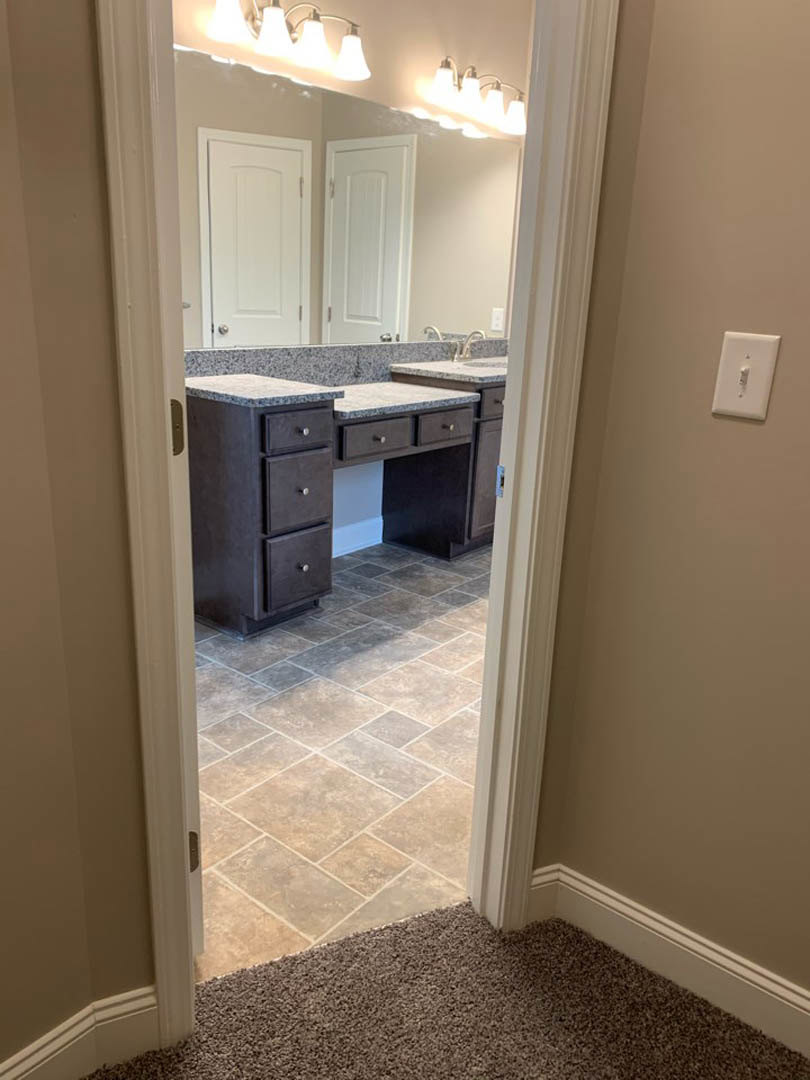 Bathroom with white tile floor, white vanity cabinet, rectangular sink, large framed mirror, silver faucet, white door with silver handle, light switch on wall, carpet visible in